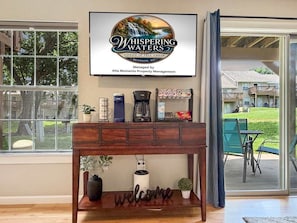 Ein Holzkonsolentisch mit einer Kaffeemaschine, einer Pflanze und einem Willkommensschild. Ein gerahmtes Schild über dem Tisch wirbt für Whispering Waters Condo in Fall Creek, verwaltet von Aha Moments Property Management.