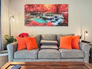 A living room with a grey sofa, orange cushions, a wooden coffee table, and a painting of a waterfall. - No stairs, clean peaceful secluded, close to strip (Branson)
