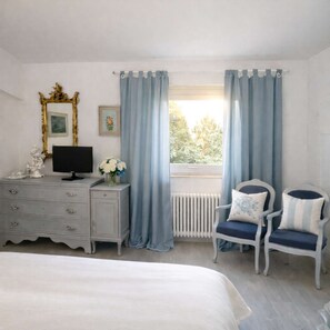 Ein Schlafzimmer mit Bett, Kommode, Fernseher, zwei Stühlen und einem Fenster mit Vorhängen.