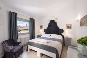 Ein Hotelzimmer mit einem großen Bett, zwei Nachttischen mit Lampen, einem Sessel, einem Fenster mit Vorhängen und Blick auf Gebäude.