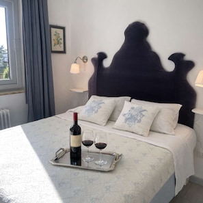 Ein Bett mit dunklem Kopfteil, weiße Kissen mit blauer Stickerei, ein Tablett mit einer Weinflasche und Gläsern sowie zwei Wandlampen.