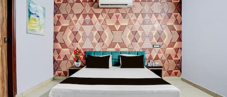 A hotel room with a geometric patterned wall, a bed with a wooden headboard, a nightstand with a lamp, and a wall-mounted air conditioner.