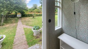 Blick auf einen Garten mit einem Kiesweg, der zu einer Tür führt, und ein weißes Interieur mit Blumen-Tapete und einer Deckenleuchte.