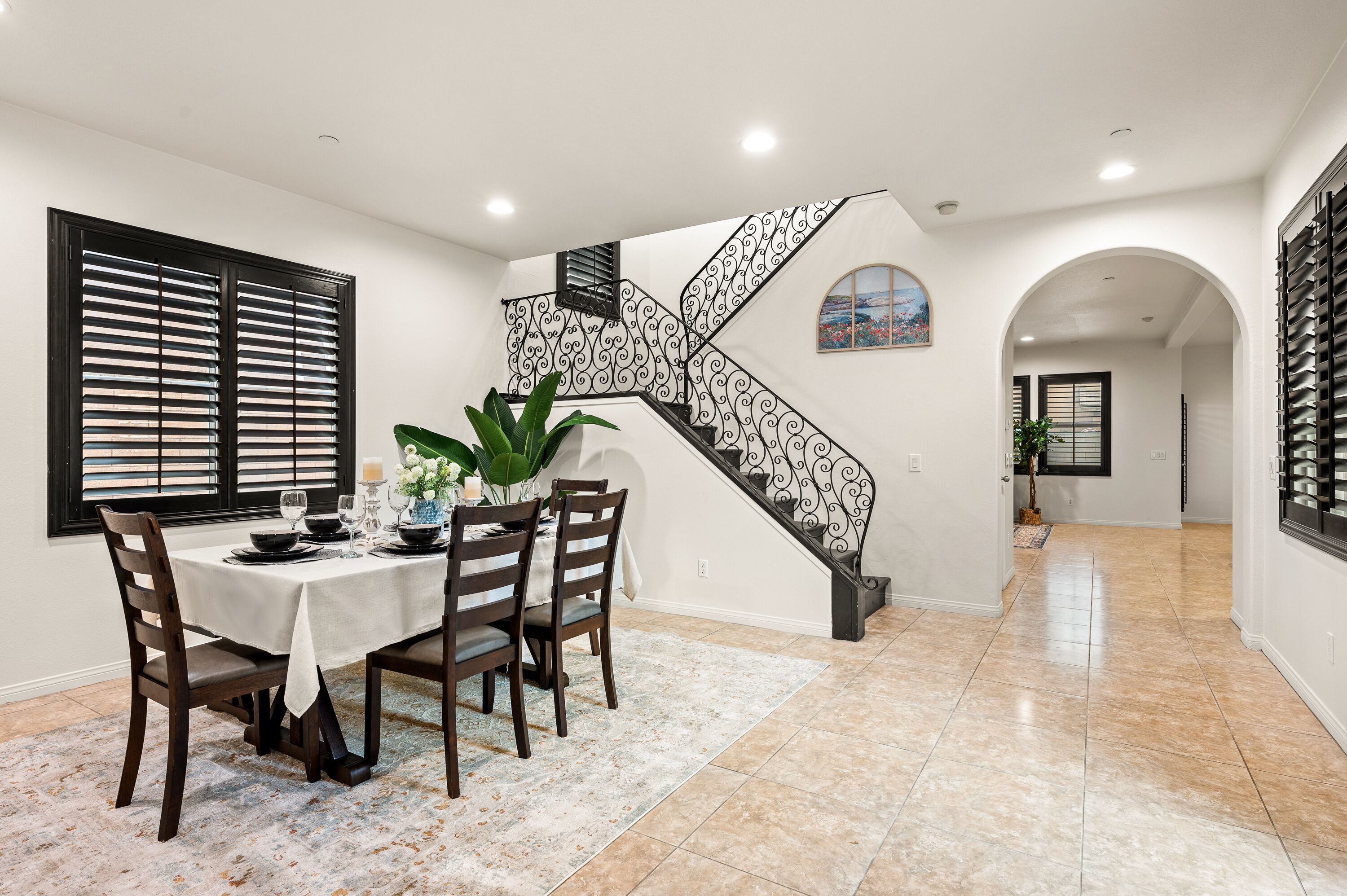 A dining area with a table set for four, a staircase with ornate railings, and a view of the adjacent room through an arched doorway.