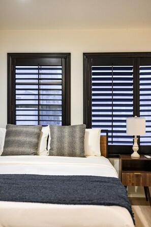 A bedroom with a bed, two pillows, a bedside table with a lamp, and two windows with blinds.