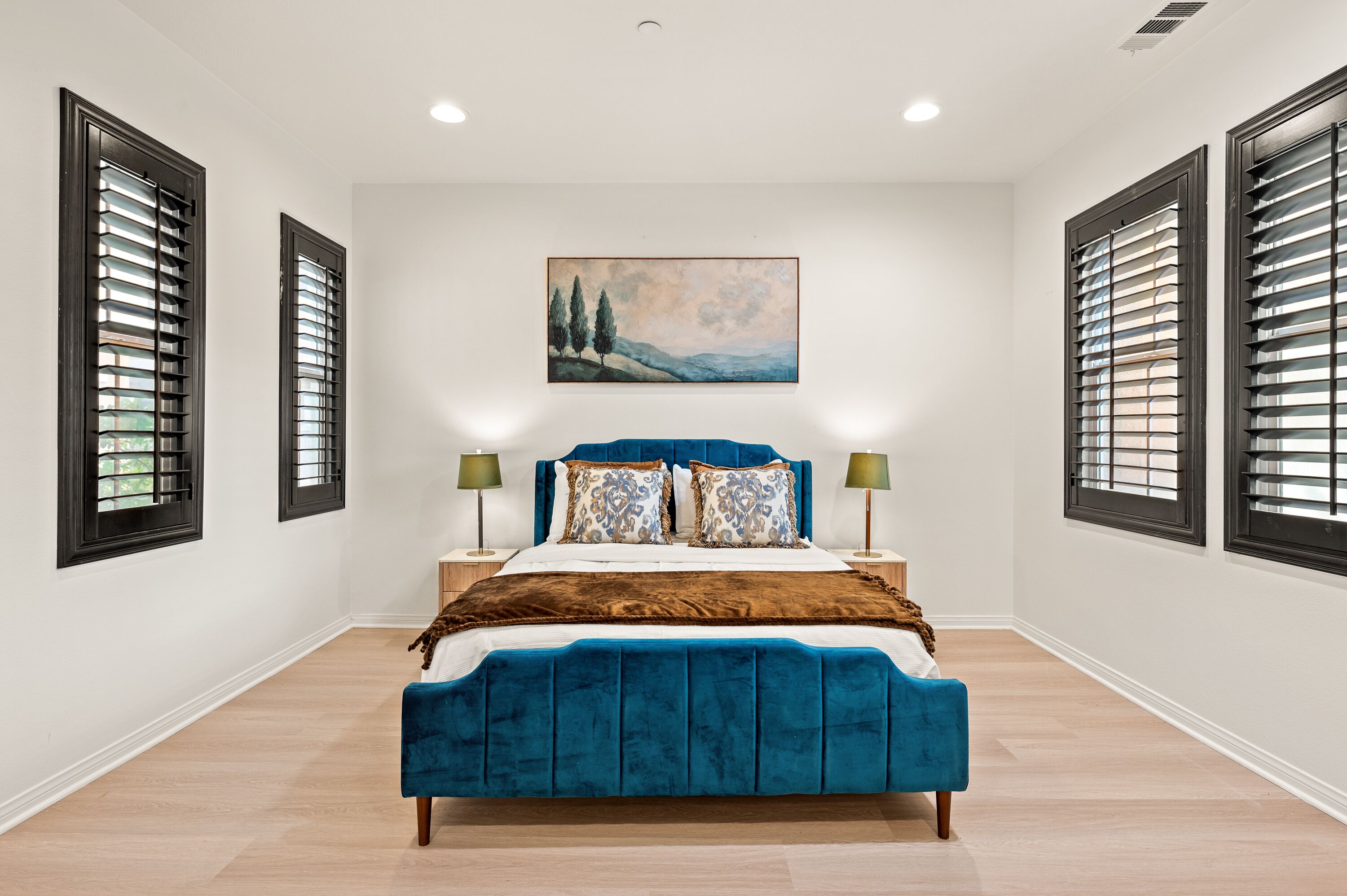 A bedroom with a large bed, two bedside tables, a painting on the wall, and a window with blinds.