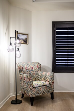 A corner room with a patterned armchair, a floor lamp, a painting on the wall, and a window with blinds.