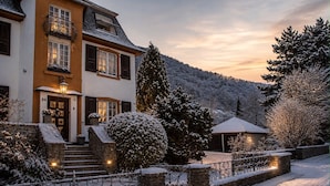 Ein zweistöckiges Haus mit einer Steintreppe, die zum Eingang führt, umgeben von schneebedeckten Bäumen und einem Berg im Hintergrund.
