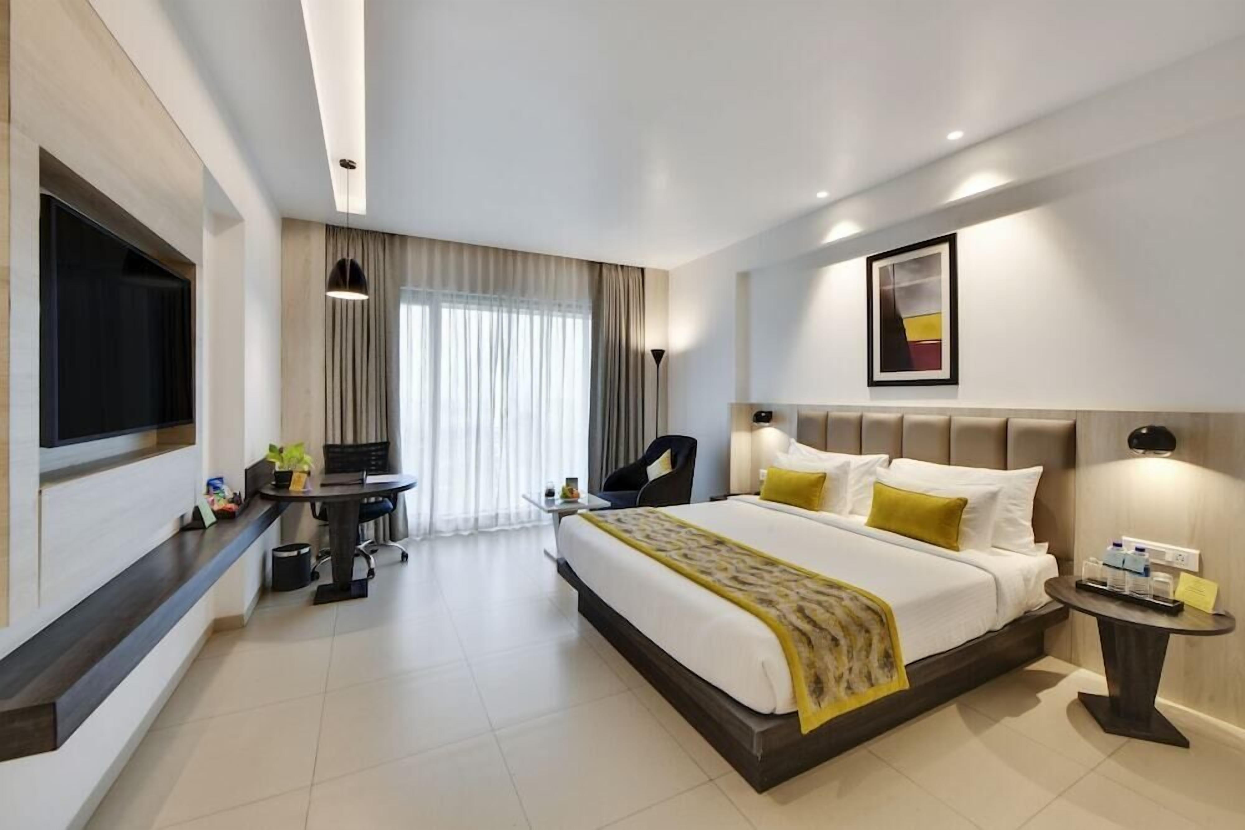 A modern hotel room with a large bed, a desk, a chair, a TV, and a bedside table.
