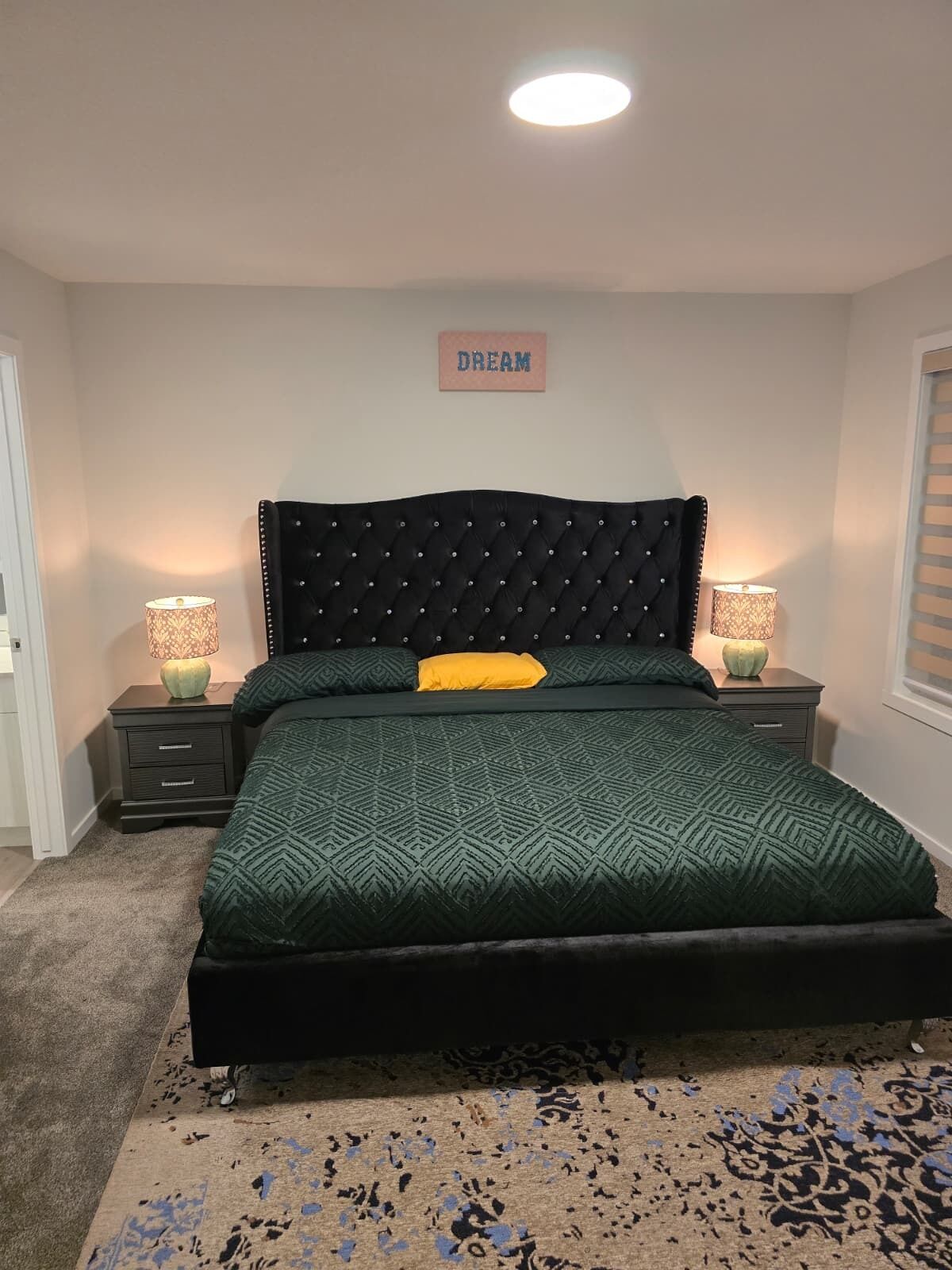 Een slaapkamer met een gestoffeerd hoofdeinde, een bedspread met een groen patroon, één geel kussen, twee bedlampen en een bordje met de tekst 'DREAM' aan de muur.