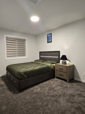 A bedroom with a bed, a nightstand, a lamp, a window with blinds, and a picture on the wall. - Modern Spacious Home - Near Airport (Northeast)