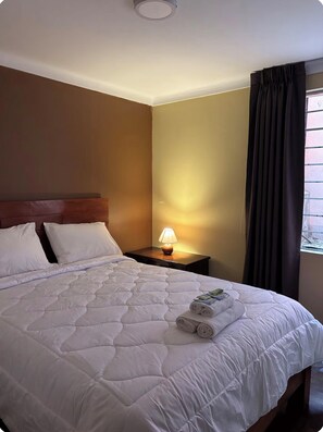 A hotel room with a large bed, a bedside table with a lamp, and a window with curtains. - Apartment in the Amauta 121 Housing Complex (Cusco)
