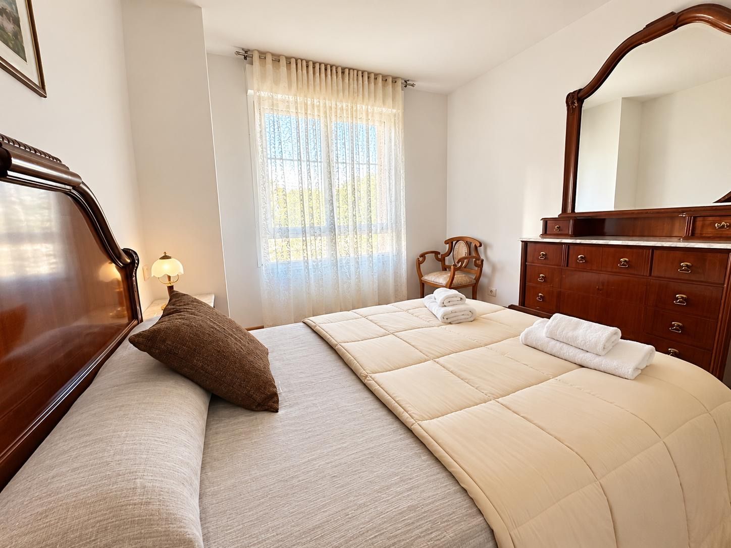 A hotel room with a bed, a wooden dresser, a chair, and a window with curtains.
