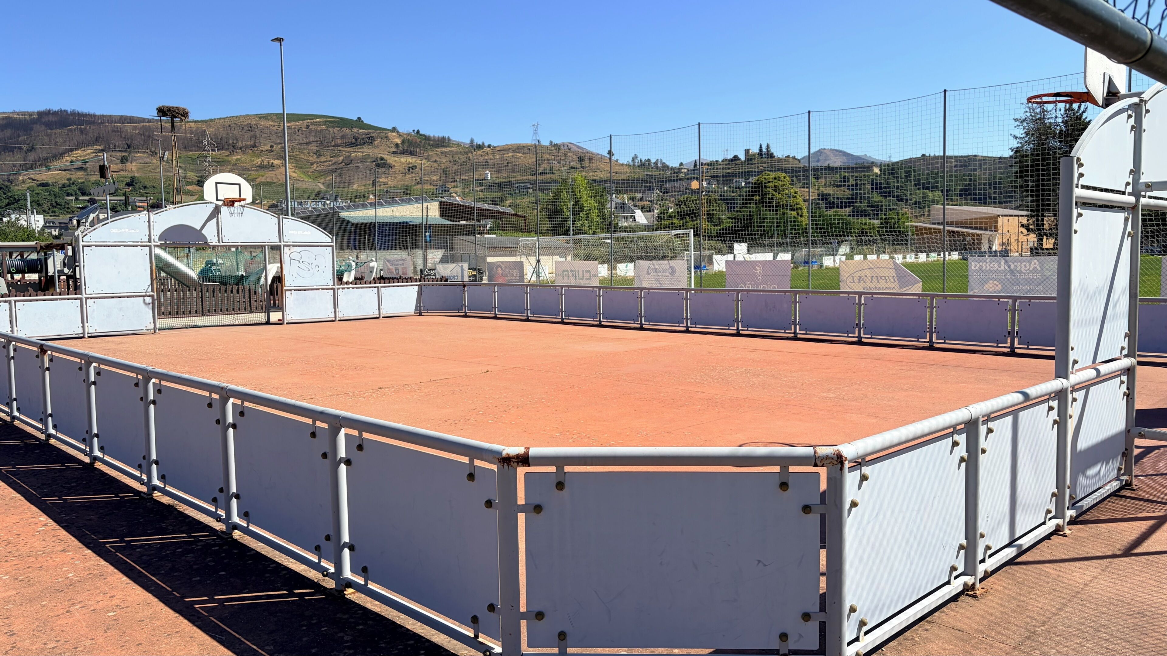 Una cancha deportiva al aire libre con un aro de baloncesto, rodeada por una valla, y una vista de un paisaje montañoso al fondo.