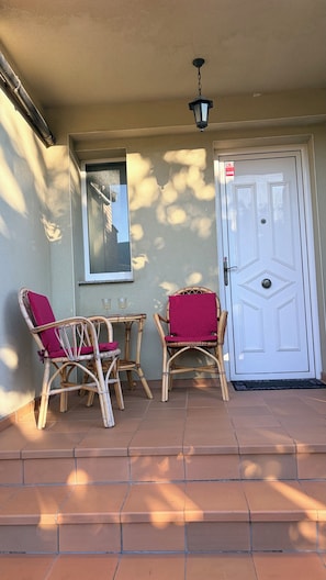 A covered patio area with two wicker chairs, a small table, and a door with a decorative glass panel.