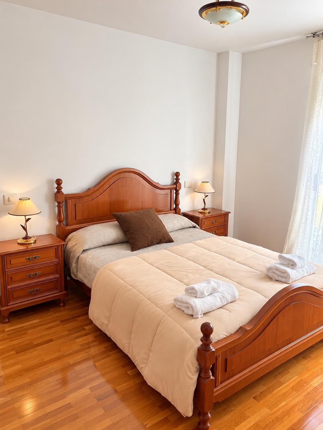 A neatly made bed with a wooden headboard, a bedside table with a lamp, and a window with curtains.