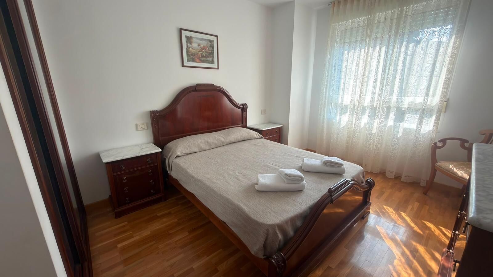 A bedroom with a wooden bed, a nightstand, a chair, and a window with curtains.