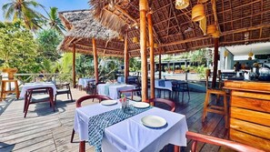 Un área de comedor al aire libre con mesas y sillas, cubierta por un techo de paja, rodeada de palmeras.
