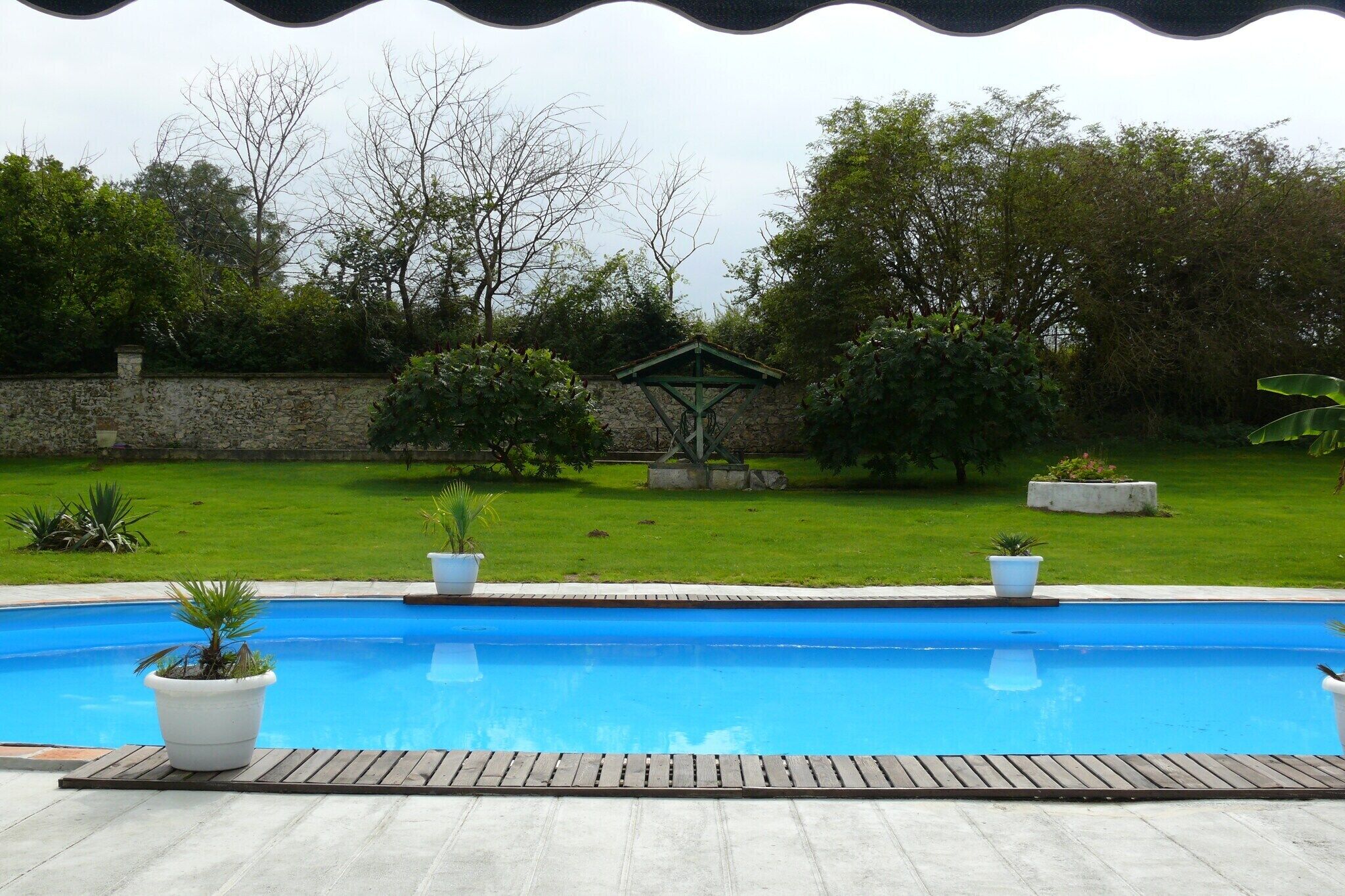 A swimming pool with a wooden deck, surrounded by a stone wall and greenery.