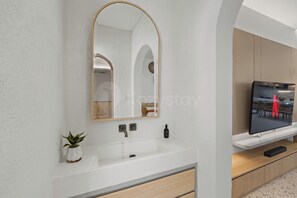 A modern bathroom with a white sink, a mirror with an arch frame, a small potted plant, and a television on a wooden shelf.