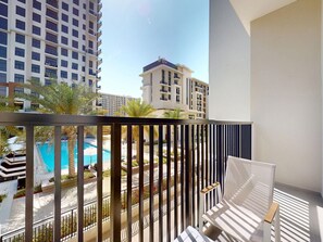 A balcony with a white chair, a pool, and high-rise buildings in the background. - Modern 2BR | Fully Furnished | Great Amenities (Dubai)