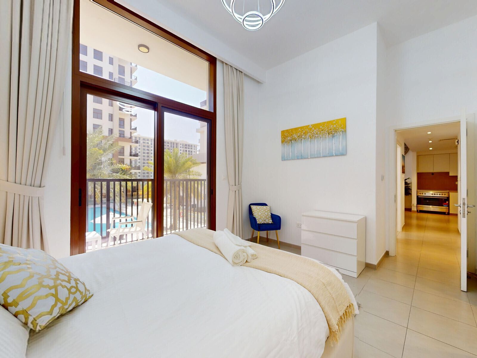A hotel room with a large bed, a view of a swimming pool, a painting on the wall, and a kitchenette.