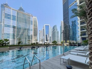 A swimming pool with lounge chairs, surrounded by high-rise buildings. - Fully Furnished 1BR | Near Metro Station | (Dubai)