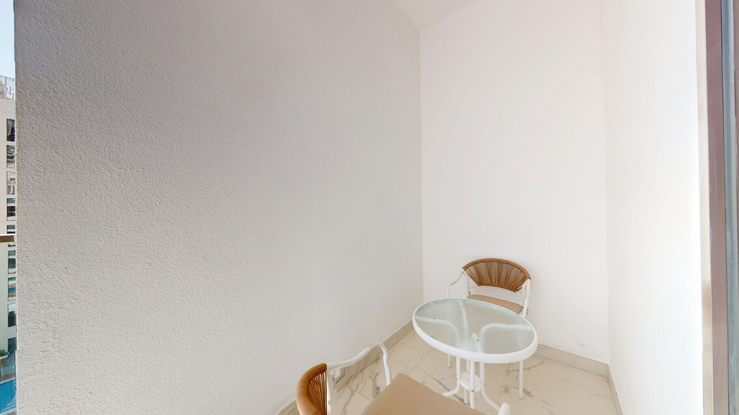 A small, enclosed balcony with a white wall, a glass table, and two wicker chairs.