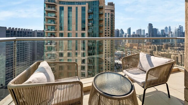 A balcony with wicker furniture and a glass table, overlooking a cityscape with the Emaar building in the background. - Luxury Downtown Living | 2BR | High Floor (Dubai)
