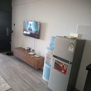 A room with a television displaying a movie, a water dispenser, a refrigerator, and a wooden bench.
