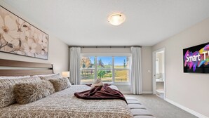 Une chambre avec un grand lit, une fenêtre donnant sur des arbres et un immeuble, et un tableau accroché au mur.