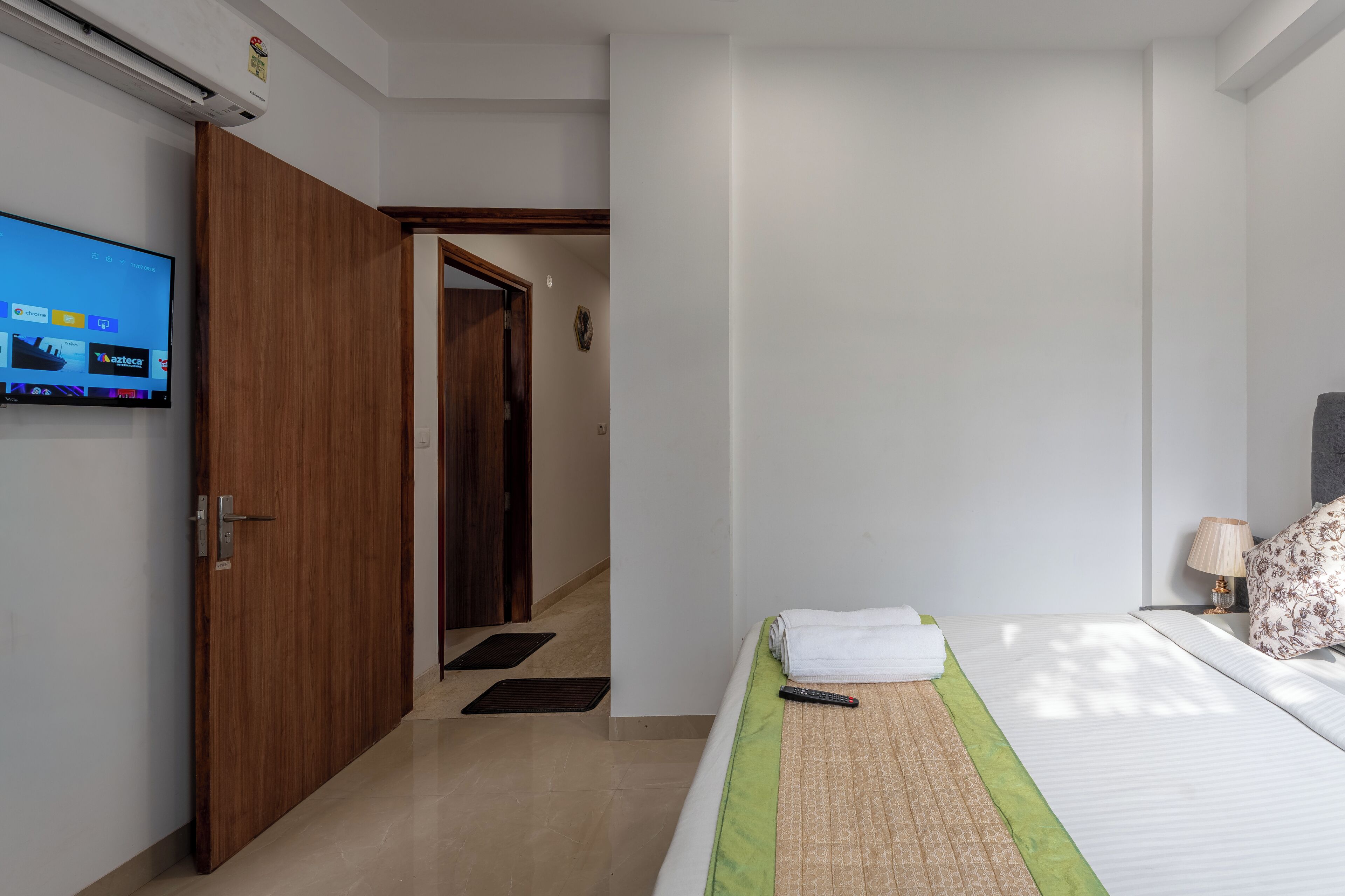 A hotel room with a bed, a TV mounted on the wall, a wooden door, and a small hallway.