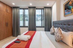 A hotel room with a large bed, grey curtains, a grey headboard, and a painting on the wall. - Divine India Service Apartment E-69 Spacious 3 Bedroom with AC & Balcony (New Delhi)