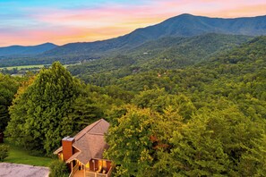 A cabin with a deck, surrounded by dense forest and mountains in the background. - Cozy Cabin + Hot Tub + Decks | Arcade + Mountain Views Near Park | Dancing Bear (Pigeon Forge)