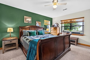Un dormitorio con cama de madera, paredes verdes, ventilador de techo, ventana con persianas y mesita de noche con lámpara.