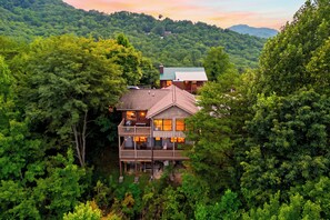 A two-story cabin with a balcony, surrounded by dense forest. - Cozy Cabin + Hot Tub + Decks | Arcade + Mountain Views Near Park | Dancing Bear (Pigeon Forge)