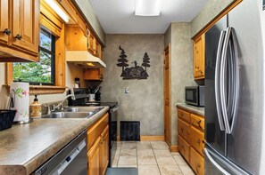 A compact kitchen with wooden cabinets, a stainless steel refrigerator, and a wall-mounted microwave. - Cozy Cabin + Hot Tub + Decks | Arcade + Mountain Views Near Park | Dancing Bear (Pigeon Forge)