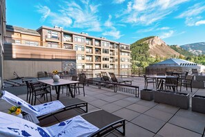 An outdoor terrace with tables and chairs, a view of a mountain, and a building with multiple balconies. - Steps to Gondola • Shared Pool + Mountain Views • Resort Access | Gondola & Gold (Avon)