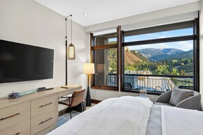 A hotel room with a bed, a desk, a TV, a lamp, and a balcony with a mountain view. - Ski Valet + Resort Pool + Hot Tubs | Valley Views | Riverfront Perch (Avon)