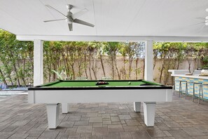 Ein Billardtisch mit Deckenventilator darüber, umgeben von einer Terrasse mit Blick auf Bäume und einem Zaun.