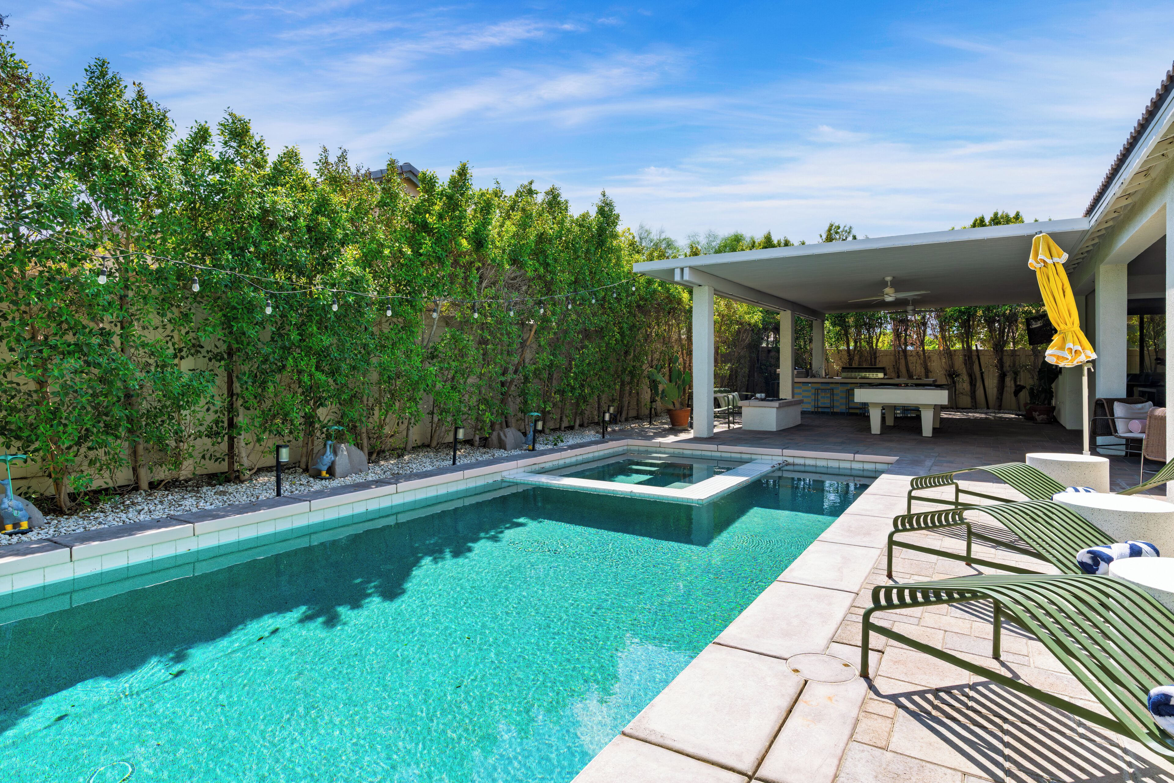 A swimming pool with a tiled deck, surrounded by lush greenery and a covered patio area.