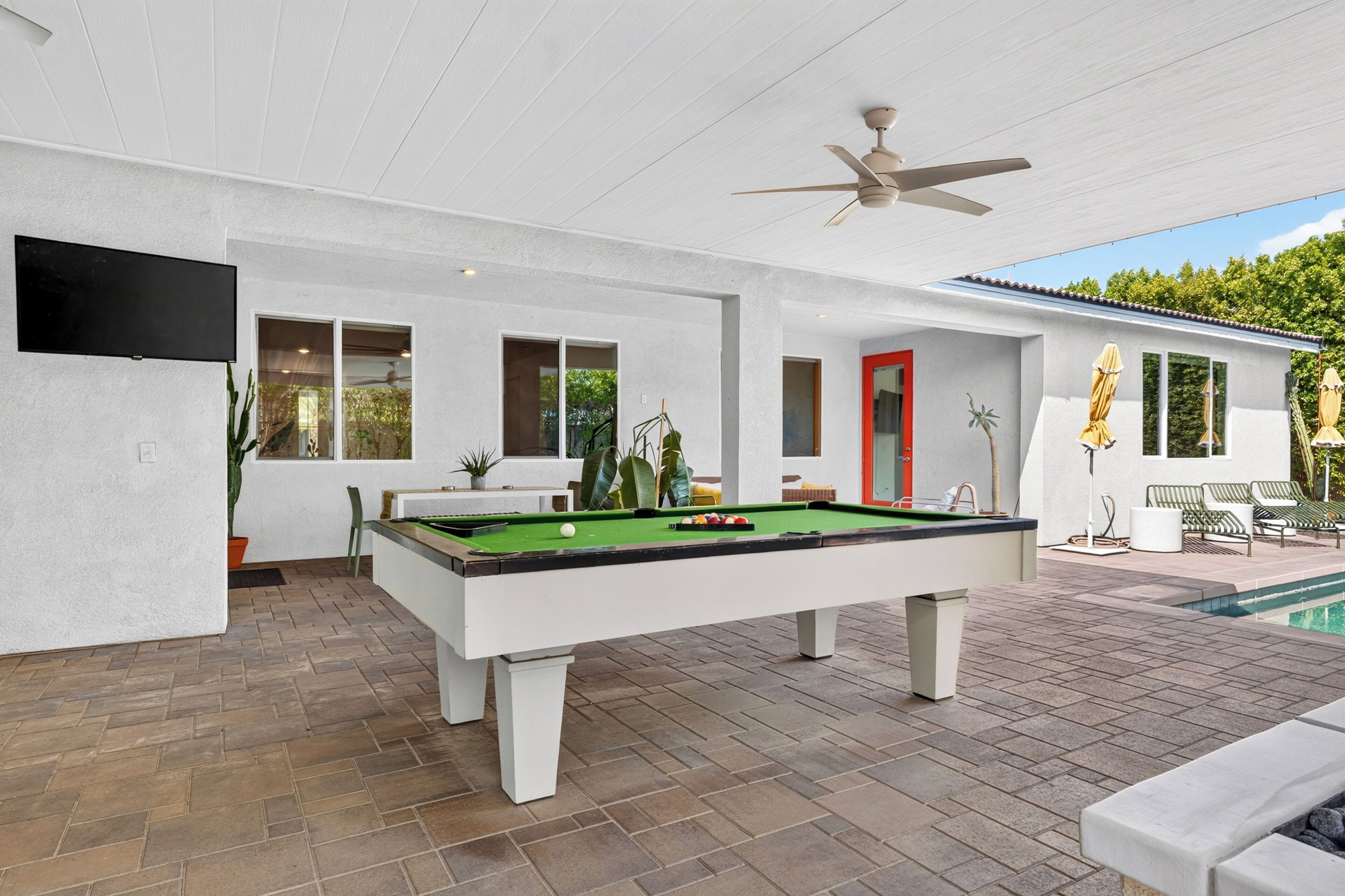 A pool table with a ceiling fan, a red door, and a poolside area with chairs and umbrellas.