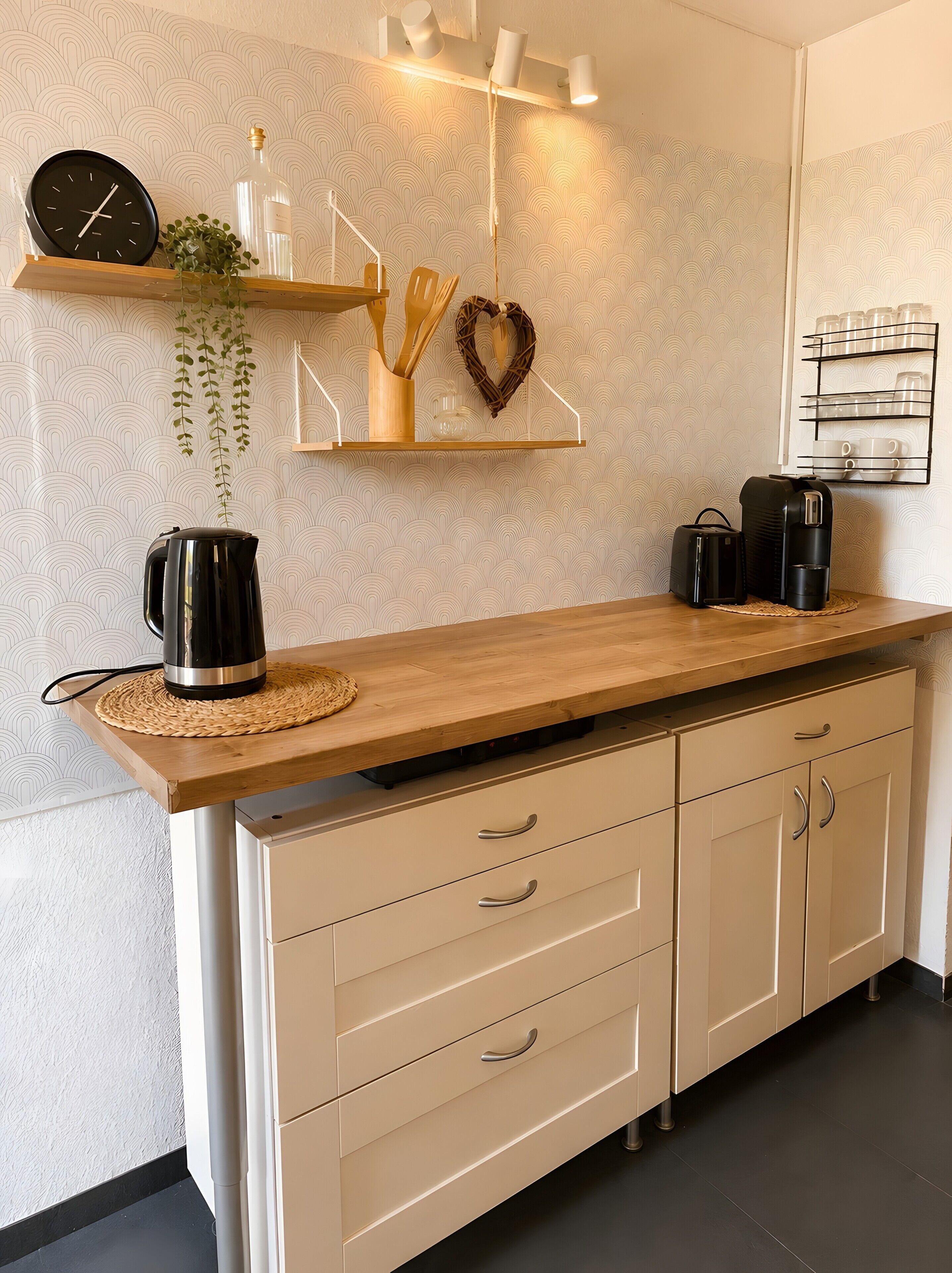 A kitchen with a wooden countertop, a kettle, a coffee maker, and a clock. There are cabinets below the countertop and a shelf with kitchenware.