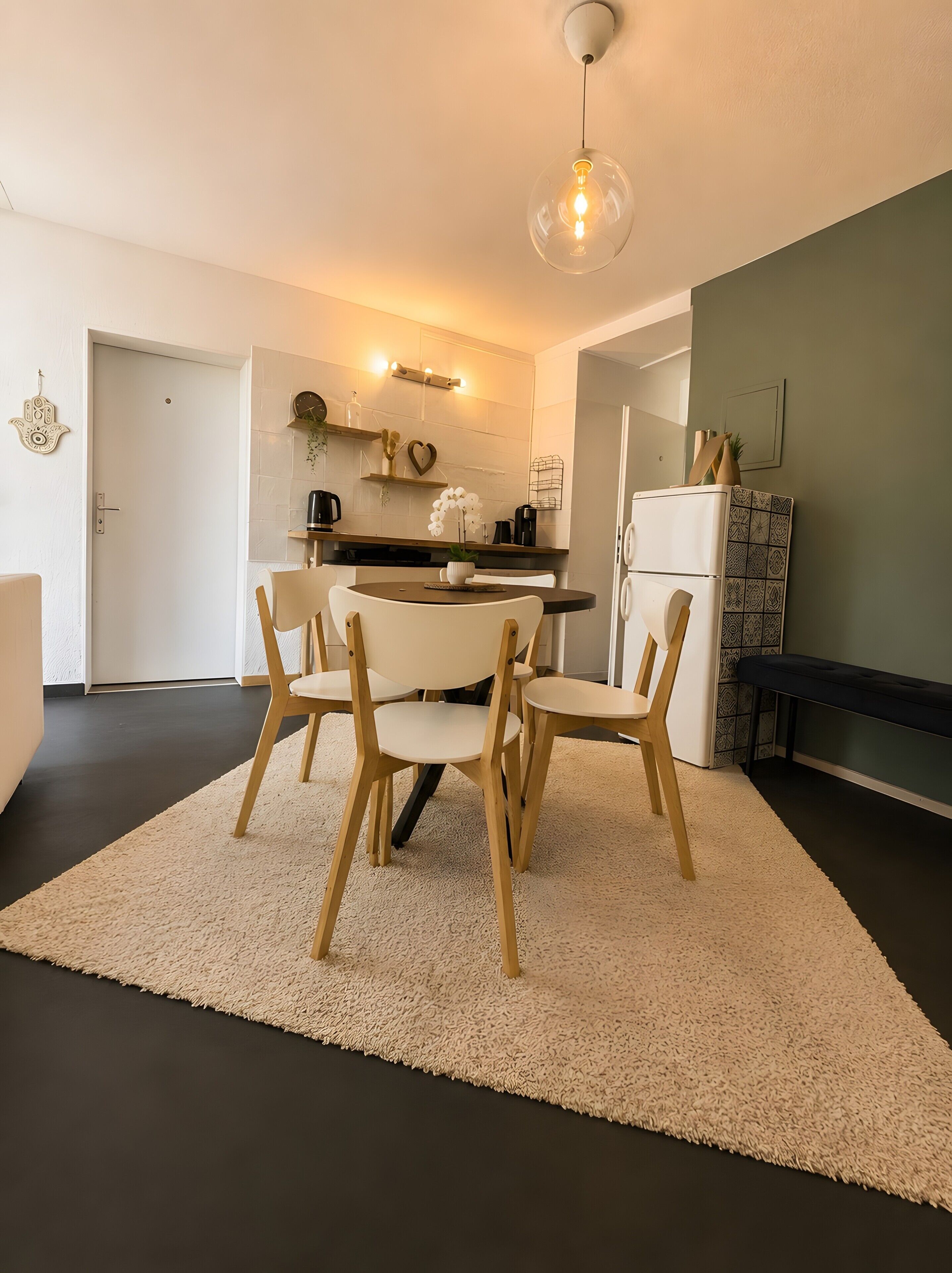 A modern kitchen with a dining area, featuring a round table with chairs, a hanging light fixture, and a refrigerator.