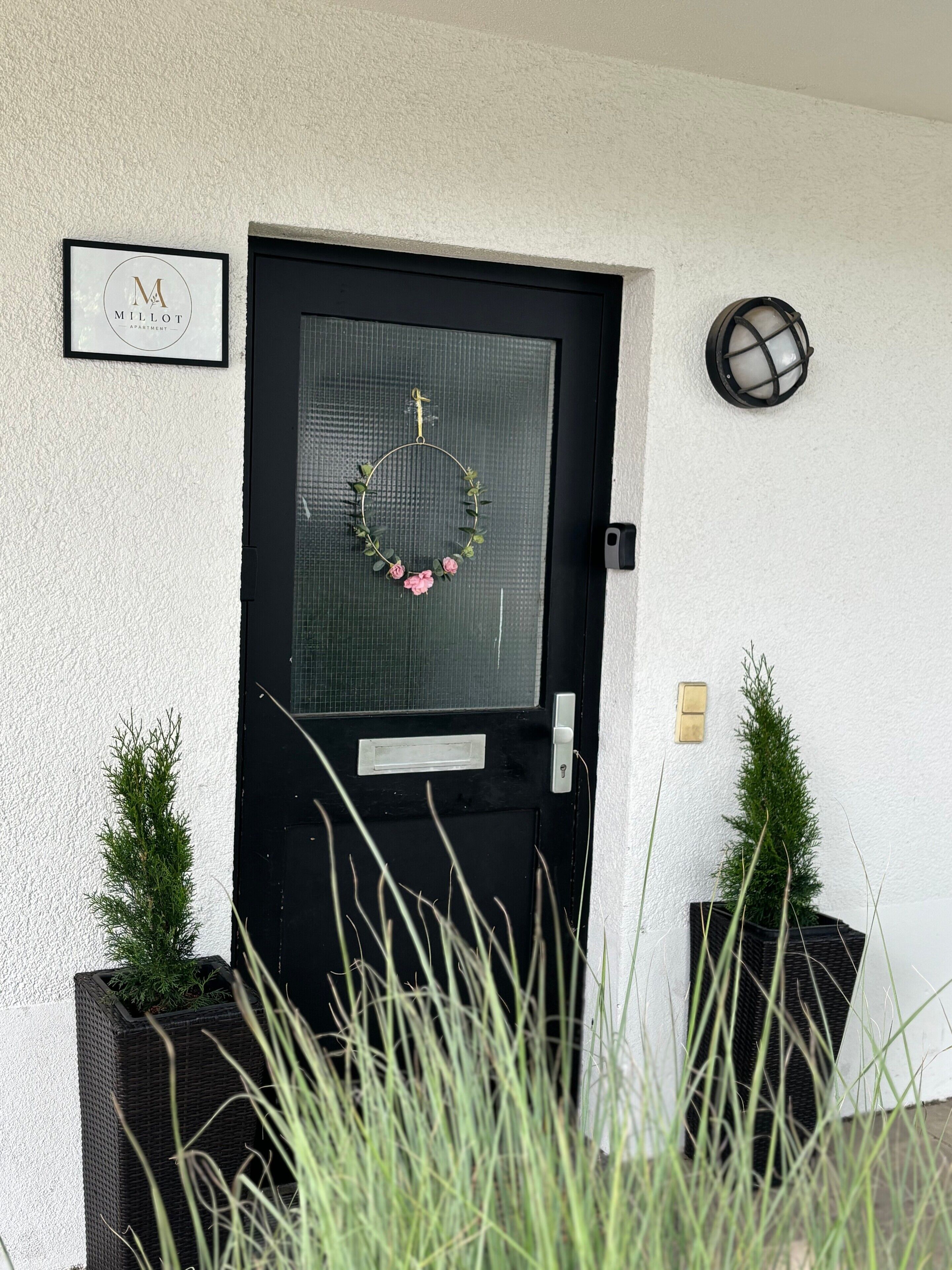 A black door with a wreath, a framed sign above, and two potted plants on either side.