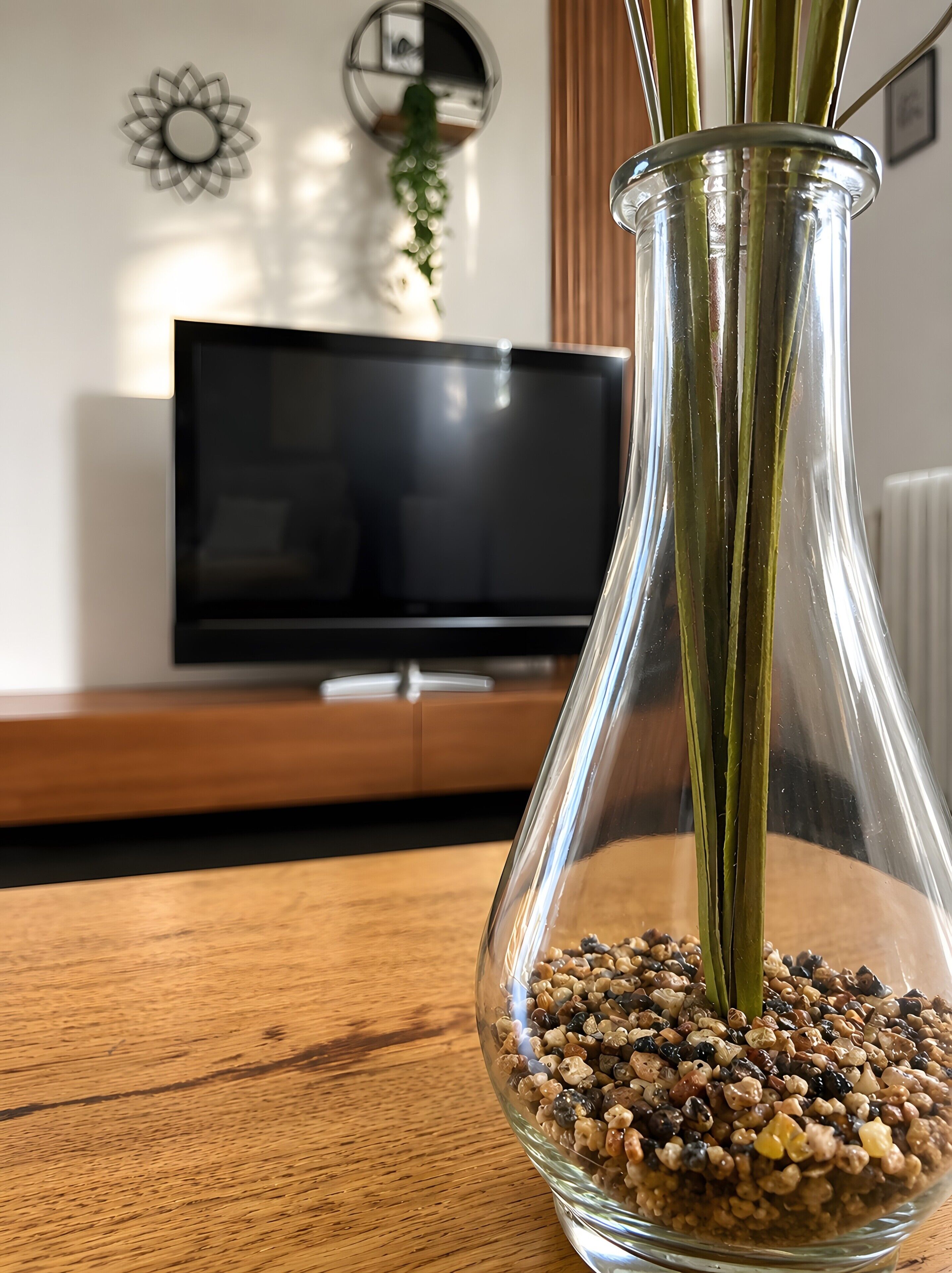Un florero de vidrio transparente con guijarros y tallos verdes sobre una superficie de madera.
