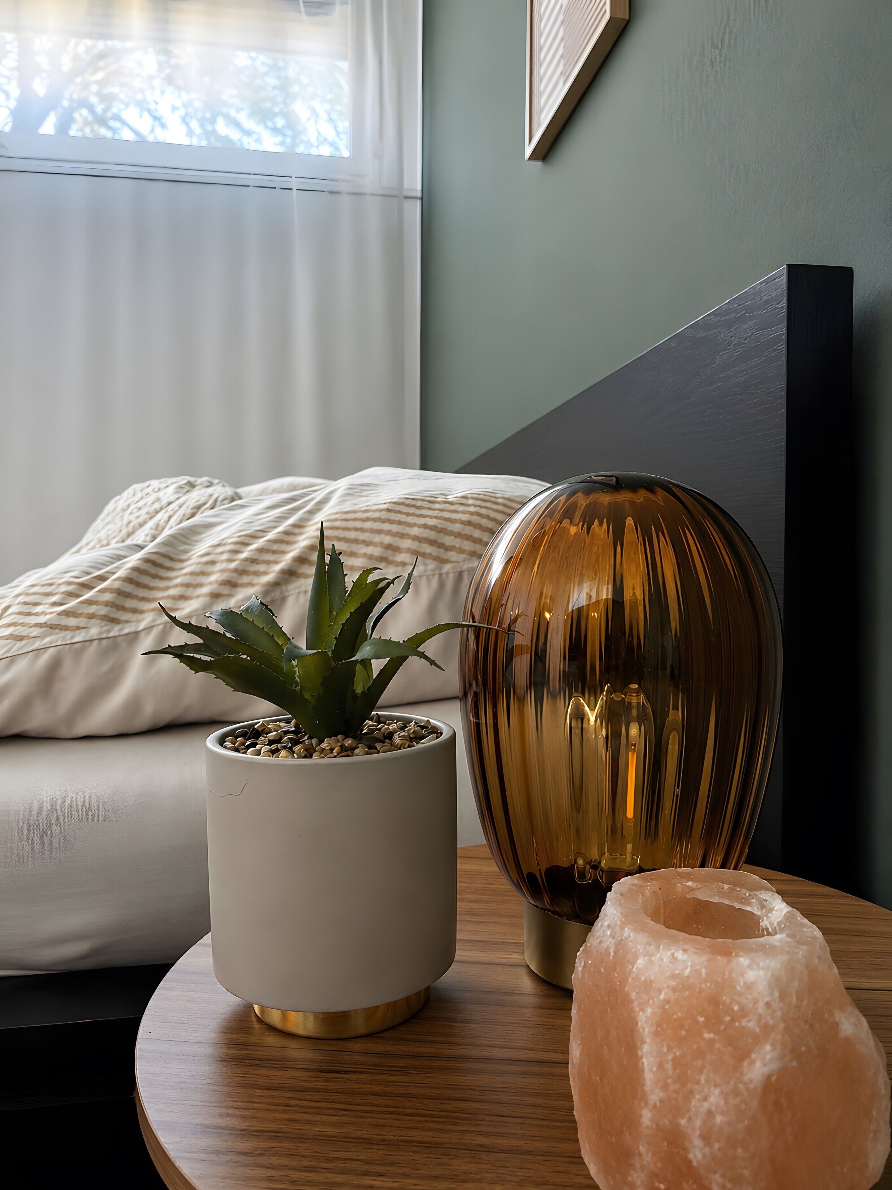 A room with a bed, a potted plant, a decorative lamp, and a salt lamp on a wooden side table.