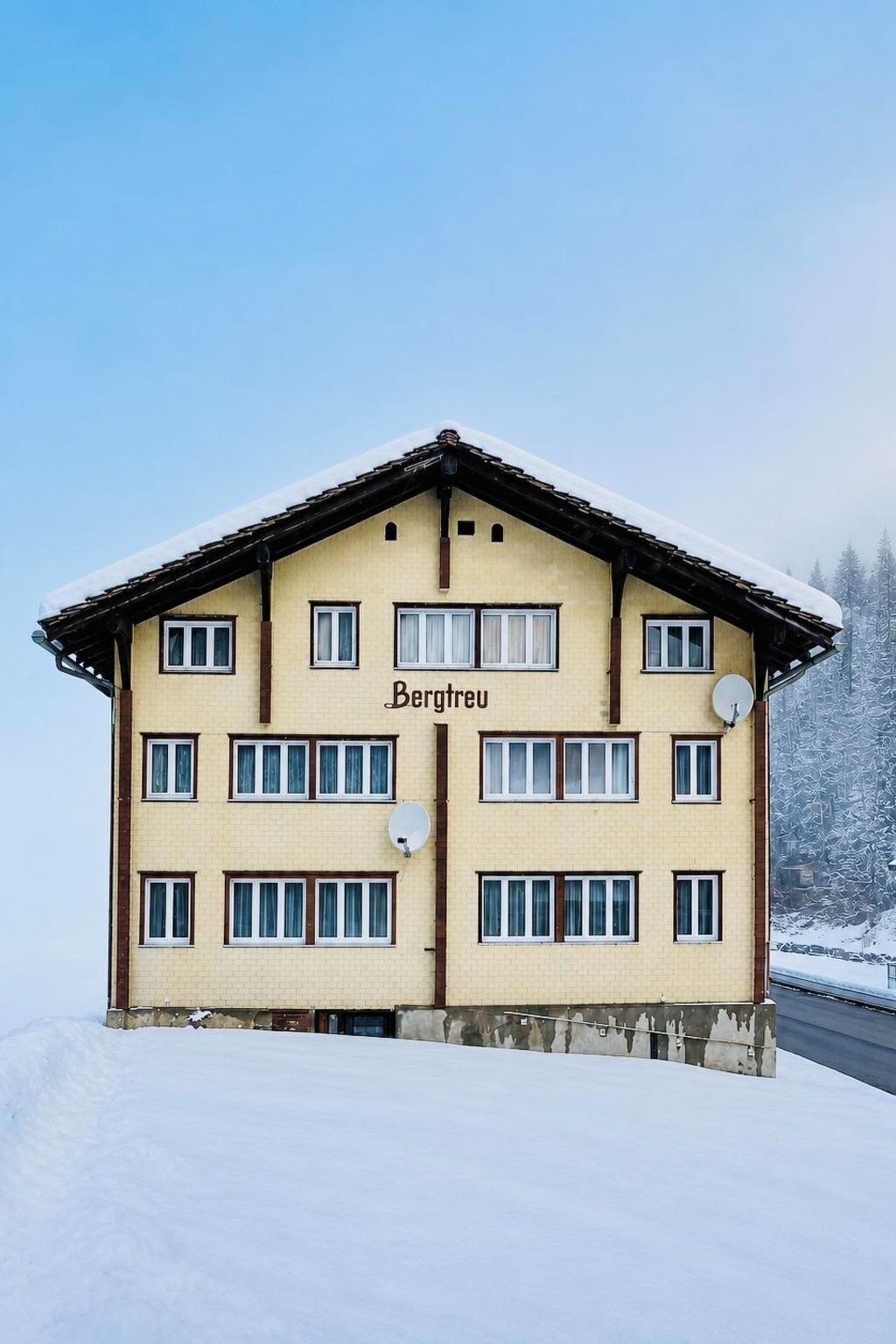 Ein mehrstöckiges Gebäude mit schneebedecktem Dach, beschriftet mit 'Bergtreu', inmitten einer verschneiten Landschaft.