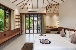 A hotel room with a large bed, a sofa, a balcony with a view of greenery, and a wooden shelf with decorative items.