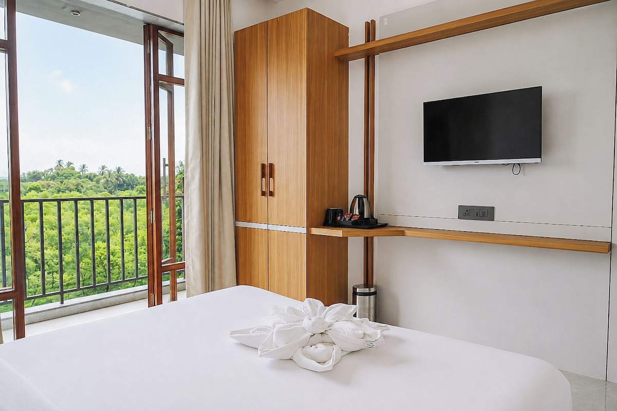 A hotel room with a bed, a TV, a wooden wardrobe, and a balcony with a view of greenery.